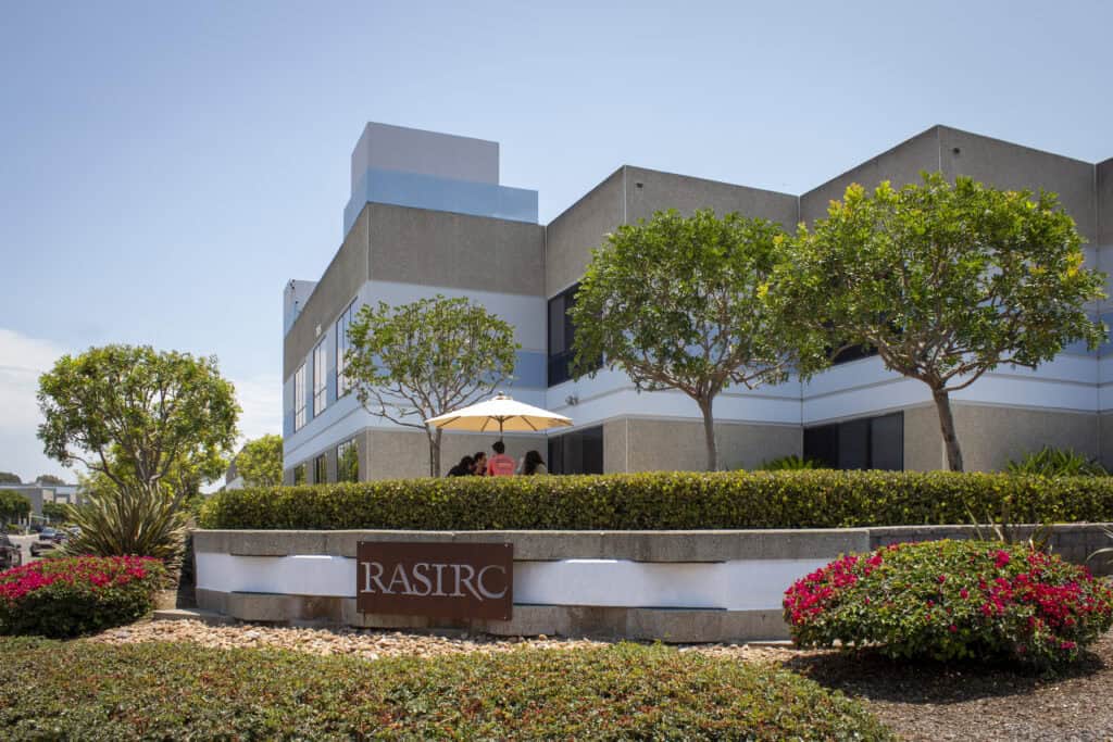 The exterior of a tan colored office building landscaped with trees and plants.